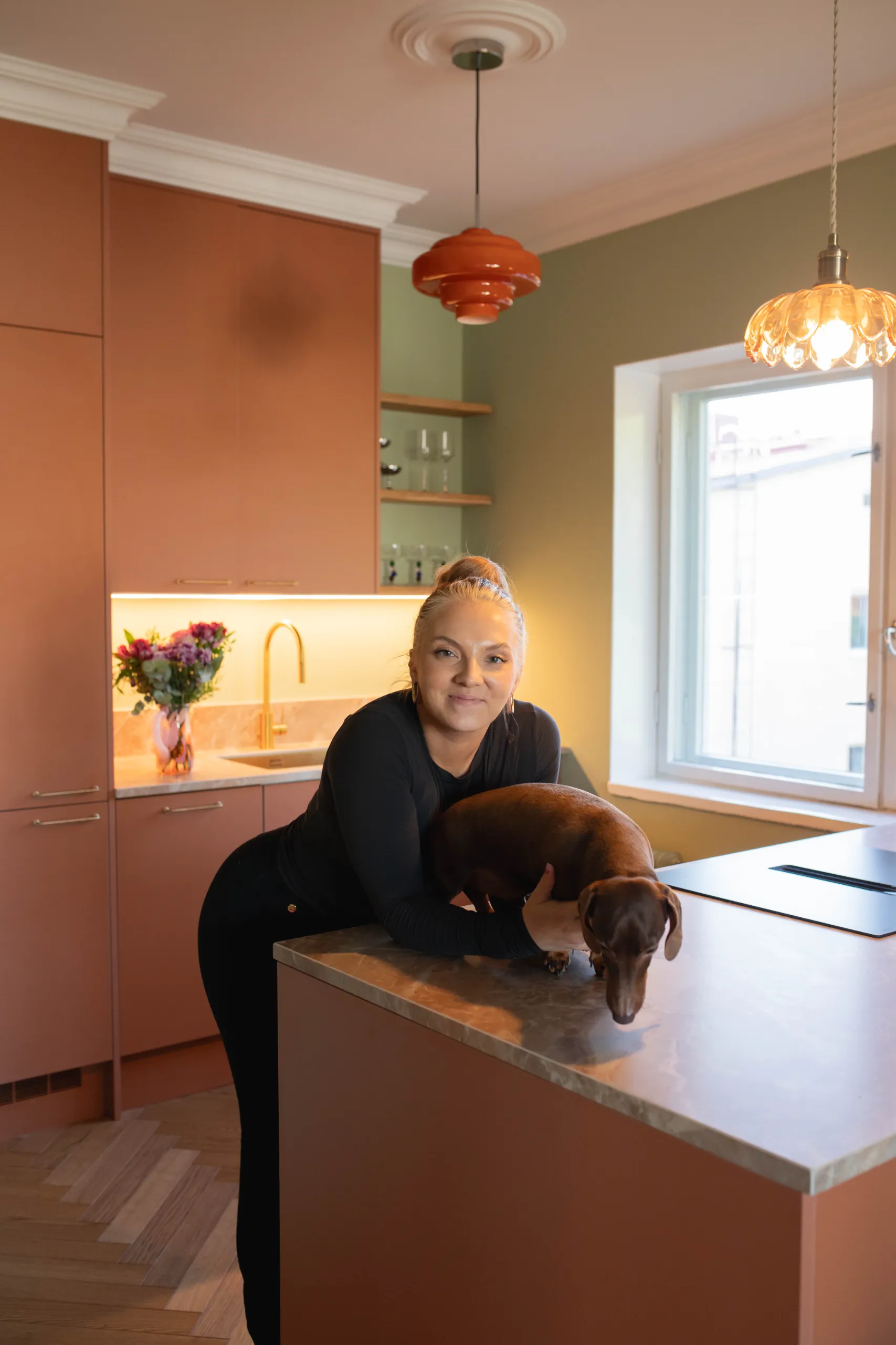 Moderni värikäs keittiö, jossa roosan sävyiset kaapistot ja salvian vihreät seinät. Katosta roikkuvat kolme valaisinta, joista keskimmäinen on punainen.